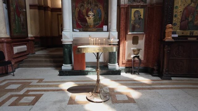 Golden candle stand with burning thin wax candles inside a solemn Orthodox church with sunbeams illuminating the stone floor, icons on the walls