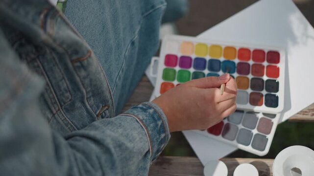 Closeup watercolor palette hand mixing tones on lap, denim jacket sleeve visible on porch, mentor tests swatches on blank paper, jars and brushes arranged, calm reflective atmosphere, experimental