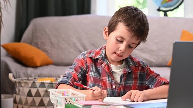Boy practices writing homework while watching educational content on laptop, creating balanced learning routine that combines technology with traditional handwriting skills