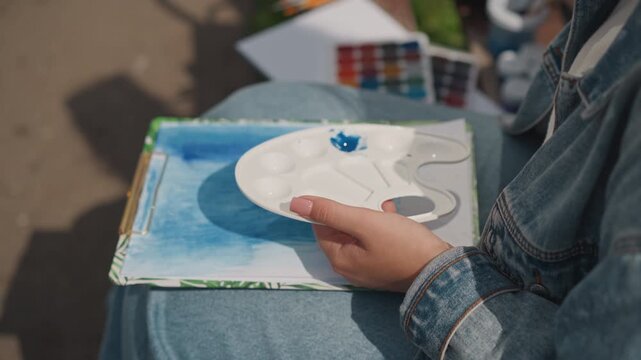White woman mixing paint on palette closeup of hand holding wells with vivid blue dab loading brush sketchbook on lap denim jacket sunlight highlights texture mindful routine preparing wash