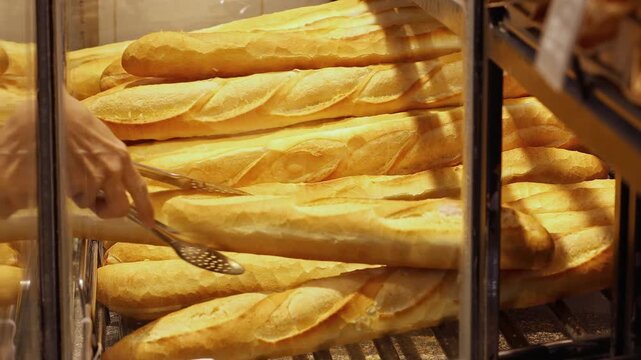 Fresh baked baguette on metal rack in bakery shop display case. Close up of golden-brown long bread loaves being handled by worker with metal tong for customer service at food market store.