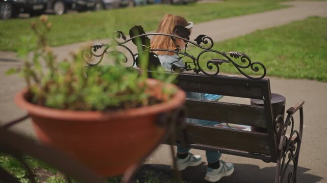 White woman sitting with back view on ornate bench with planter in foreground, contemplative pause after painting, park pathway and soft bokeh, denim and ponytail, serene afternoon and peaceful