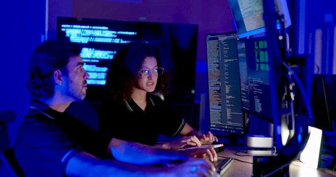 Adult male and female technology professionals type at computer workstations, review source code on multiple screens and collaborate on cybersecurity analysis in modern digital operations office.