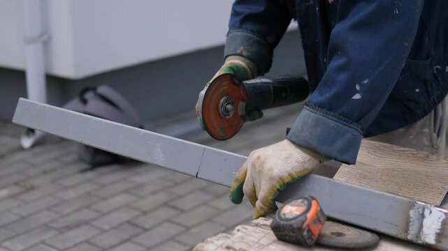 Close side view shows worker in blue workwear using handheld grinder on steel beam, with bright sparks around cutting point, worn gloves, tape measure, wooden support, soft gray light, and