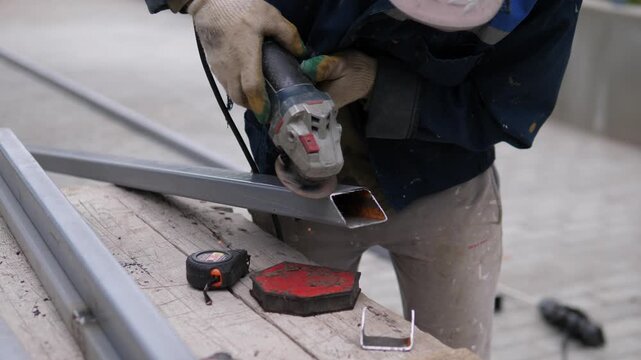 Close-up shows construction worker cutting or shaping hollow metal beam with handheld angle grinder on weathered wooden support, gloves and workwear visible, sparks flying, muted gray background