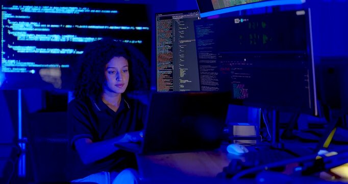 Adult female technology professional types on laptop, watches source code across monitors and shifts attention between screens during cybersecurity monitoring in modern digital operations office.