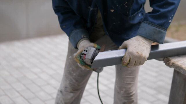 Worker in blue work jacket grinds rectangular metal tube outdoors with handheld tool, worn gloves, bright sparks, and focused repair work at contact point in industrial setting, with worker, grinder