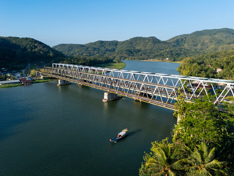 Aerial view of Jembatan Kereta Api Sungai Serayu Rawalo crossing the Serayu River with a boat, surrounded by lush hills in Kecamatan Rawalo, Jawa Tengah, Indonesia.