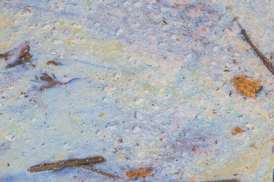 Mottled film of pollutants spreads across water in muted blues and rusty spots; organic swirls and bubble-like textures convey environmental contamination