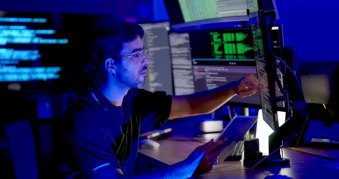 Adult male cybersecurity analyst holds tablet, points toward code monitor, checks digital system information, and reviews network security data during secure technology operations at workstation.