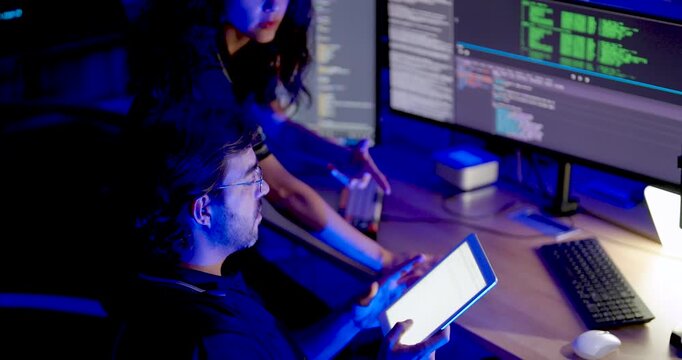 Adult male and female technology professionals review tablet and source code on monitors, pointing toward computer screens while collaborating on cybersecurity analysis in digital operations office.