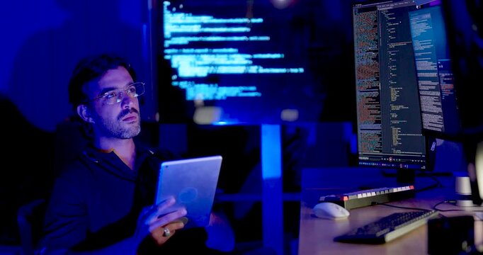 Adult Asian male cybersecurity analyst holds tablet, looks toward multiple code monitors, and gestures at screen while reviewing digital systems during network security monitoring and data analysis.