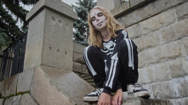 Portrait of Caucasian teenager girl wearing skeleton makeup and costume crouching on stone stairs, shot from low angle. Clip is perfect for Halloween marketing and retail campaigns