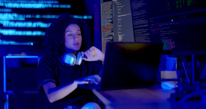 Adult female technology professional with headset around neck types on laptop, checks source code across monitors and gestures while reviewing cybersecurity data during system monitoring in office.