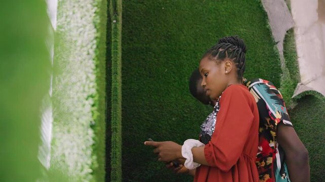 Couple checking phone near green wall her fingers scroll screen him leaning close playful debate over plans patterned shirt red dress braided hair urban backdrop casual date energy candid exchange
