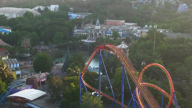Aerial view of a thrilling roller coaster with people riding through loops and drops in a vibrant amusement park, surrounded by lush green trees and other attractions during a beautiful sunset