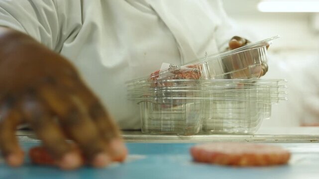 Factory worker in white coat hand packs raw beef burger patties into clear plastic trays on a moving conveyor.