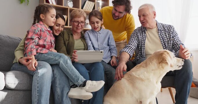 Family parents children grandparents with tablet at home. Smiling group gathers on a couch, learning online. A calm dog rests as relatives connect together. Family bonding with tech at home.