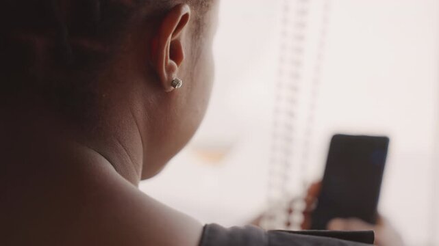 Lady examines her phone before gathering. Woman with braided hair waits quietly for her appointment. Black woman softly reviews her messages through window glow before her upcoming engagement