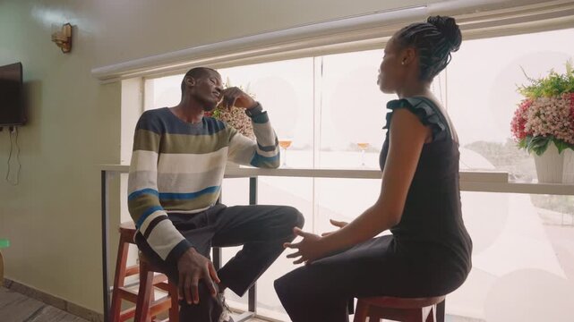 Black couple seated by sunlit window on cozy date woman gestures passionately while man sips drink, flowers in vase on sill, bar stools and soft interior light create warm atmosphere, candid emotional