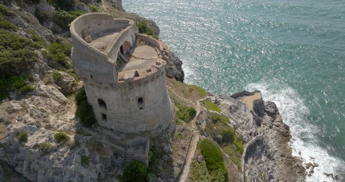 Aerial view of an ancient stone watchtower standing on a rocky cliff overlooking shimmering deep blue sea waters with gentle foam along the jagged, steep coastline in a sunny summer morning.