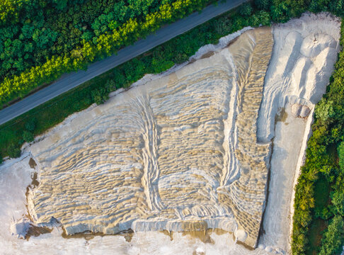 Aerial view of CCB Granulats Gaurain quarry showing textured sand and gravel piles surrounded by dense forest and a road in Tournai, Wallonia, Belgium.