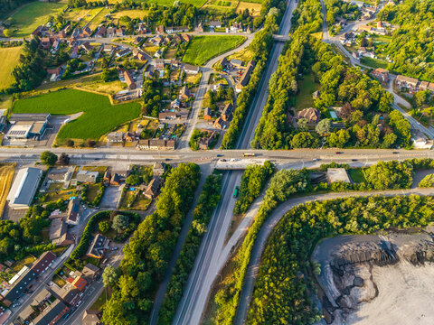 Aerial view of the CCB Granulats Gaurain quarry site adjacent to a highway intersection with surrounding residential houses, green fields, and dense trees in Tournai, Wallonia, Belgium.