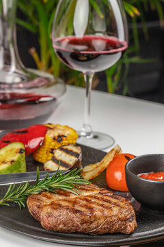 Grilled steak served on a black plate with assorted vegetables, red wine in a glass, and a decanter in the background on a white table