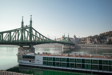 Naklejka premium Liberty Bridge steel truss structure over the Danube River in Budapest, Hungary, with a river cruise ship docked below and Gell rt Hill visible in the background.