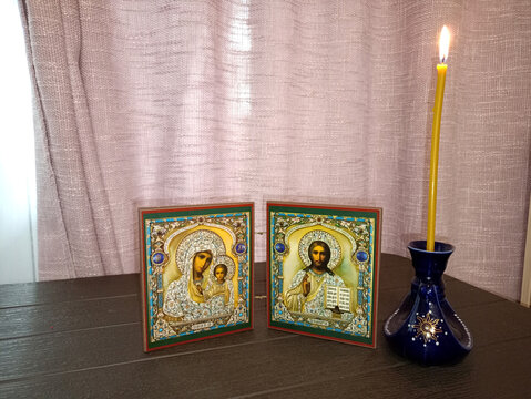 Moscow, Russia - 07.22.2026: A burning wax candle and a pair of Orthodox icons depicting the Blessed Virgin Mary (Our Lady of Kazan) and Jesus Christ (Savior, Almighty) on a table - horizontal photo, 