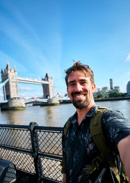 Vertical. Smiling male tourist with a backpack takes a wide-angle selfie beside the River Thames with Tower Bridge in the background on a bright sunny day in London.