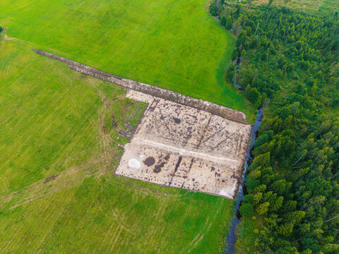 Aerial view of a rectangular patch of cleared soil surrounded by vibrant green fields and a dense forest with a small stream in Valgunde Parish, Jelgava Municipality, Latvia.