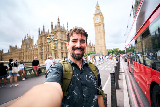 Smiling male tourist with a backpack takes a wide-angle selfie beside Big Ben and the Houses of Parliament in London, with a red double-decker bus passing nearby.