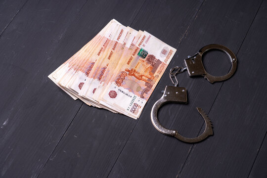 Stack of Russian ruble banknotes lies on a dark wooden surface.\nMetal handcuffs rest beside the cash.\nThis image connects money with law enforcement.\nUse it for financial crime, corruption, or lega
