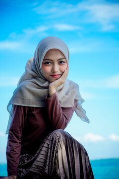Three-quarter outdoor portrait of a young Asian Muslim woman wearing a beige hijab and long-sleeved top, sitting gracefully on concrete breakwater rocks with her chin resting gently on her hand. 