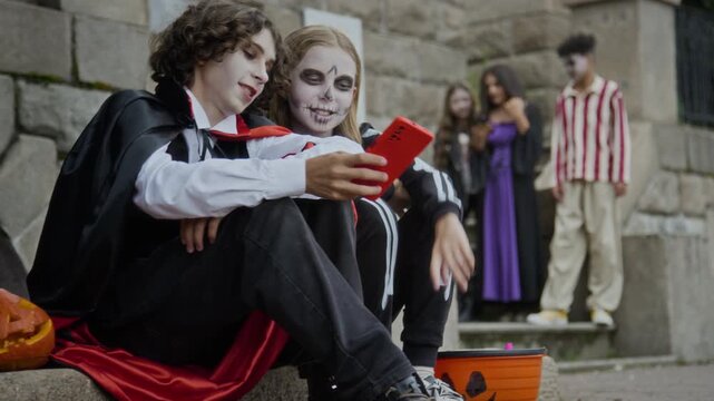 Caucasian teenagers in Halloween costumes sitting on stone steps and looking at red smartphone. Footage is ideal for seasonal holiday marketing campaigns and mobile application advertisements