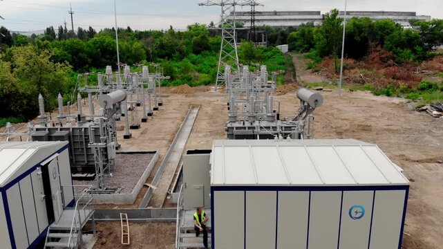 Aerial drone view of an electrical substation under construction in Sumy, Ukraine. High-voltage transformers, switchgear, transmission equipment, utility buildings, workers, and a yellow excavator