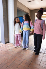 Fototapeta premium African American teenage girls holding hands and walking in school hall with backpacks near lockers