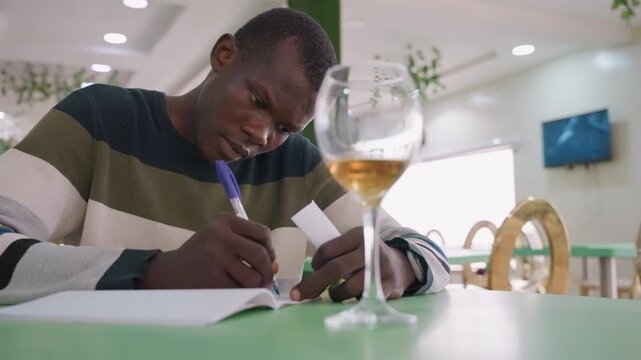 Man writing love note in cafe, wine glass foreground, focused expression, striped sweater, green table, handwritten message in notebook, soft bistro lighting, intimate romantic atmosphere.