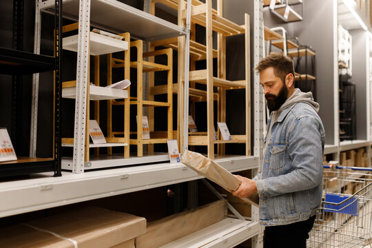 Bearded man choosing furniture box near wooden shelves in department home improvement store flat pack cardboard box shopping cart retail hypermarket warehouse commercial business buyer male consumer