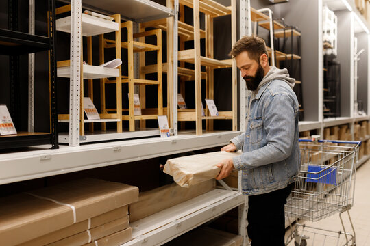 Bearded man choosing furniture box near wooden shelves in department home improvement store flat pack cardboard box shopping cart retail hypermarket warehouse commercial business buyer male consumer