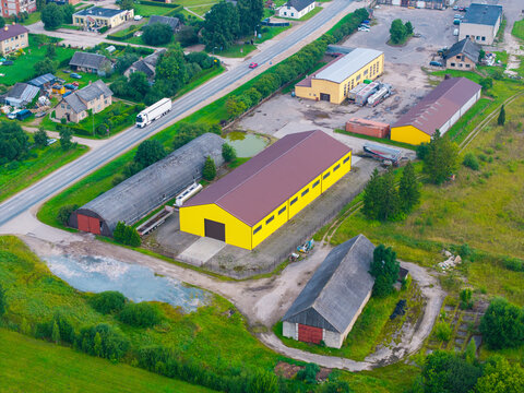 Aerial view of an industrial complex featuring a prominent yellow warehouse and arched storage buildings surrounded by green fields and a road in Jaunberze, Dobele Municipality, Latvia.