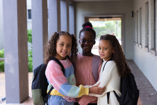 Diverse female students hugging and smiling in school walkway, wearing backpacks and sweaters