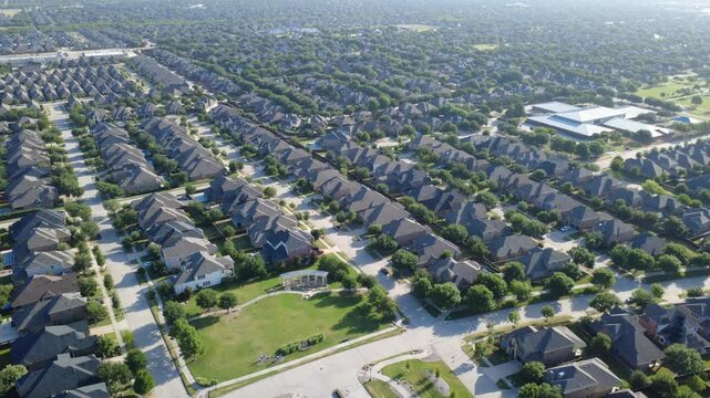 Sprawling public school building borders dense residential blocks in Denton County. High drone angle captures two story brick houses, dark shingle roofs, backyard pools, park lawns, Frisco, TX