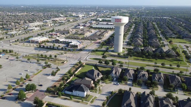 Commercial parking bays border multi lane thoroughfares near Phillips Creek Ranch in North Texas region. Elevated perspective details civic utility tower, retail plazas, home rows in Frisco, TX