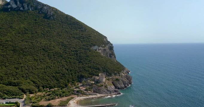 Aerial view from Sabaudia of Mount Circeo rising dramatically above a canal, sandy beach, and historic watchtower perched on rocky cliffs along the deep blue sea. Mediterranean panorama.