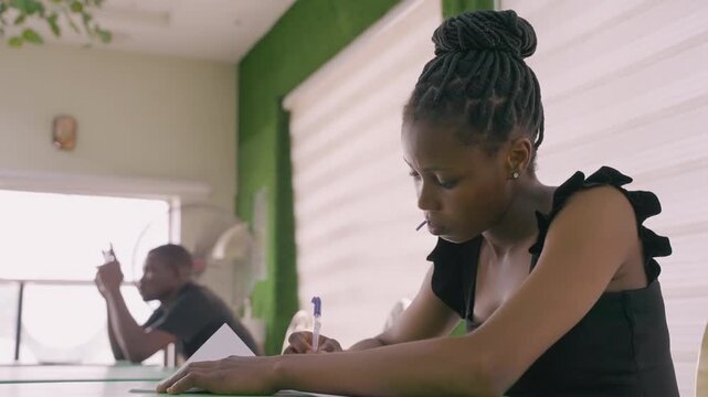 Cafe date couple at table woman in black top writing notes while partner scrolls phone by window soft sunlight washes green interior pen notebook coffee glass thoughtful quiet intimacy candid moment