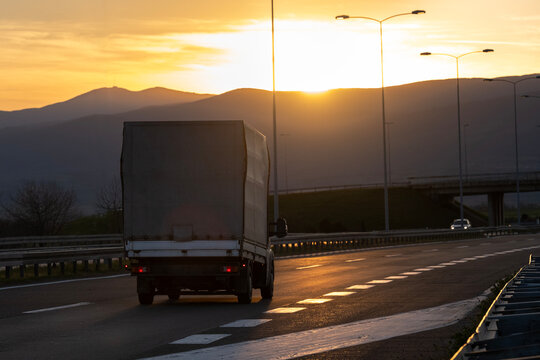 Silhouette of a curtain-side delivery truck on a highway at sunset with mountains and street lights in the background