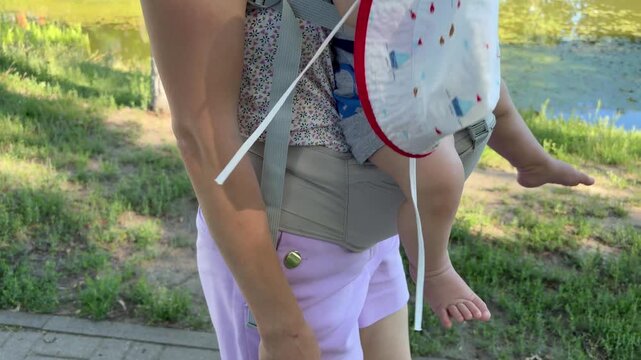 A mother walking in a park carrying her baby in a front-facing ergonomic carrier. Modern parenting and family outdoor activities