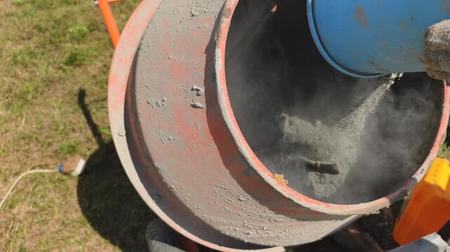 Dry cement powder enters rotating drum during mortar recipe mixing. Water pours inside electric concrete mixer, creating perfect mortar recipe with required strength for construction work.